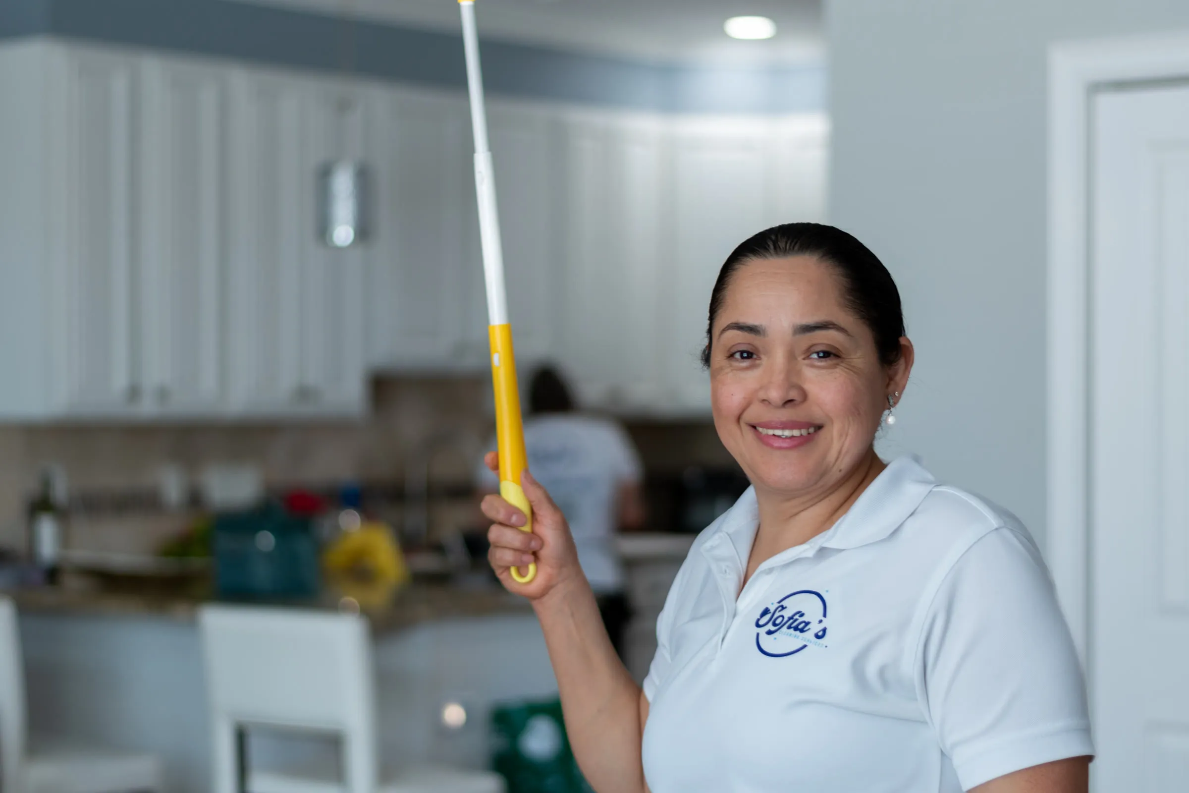 Sofia of Sofia’s Cleaning Services, in a branded polo shirt, holding a duster in a freshly cleaned kitchen